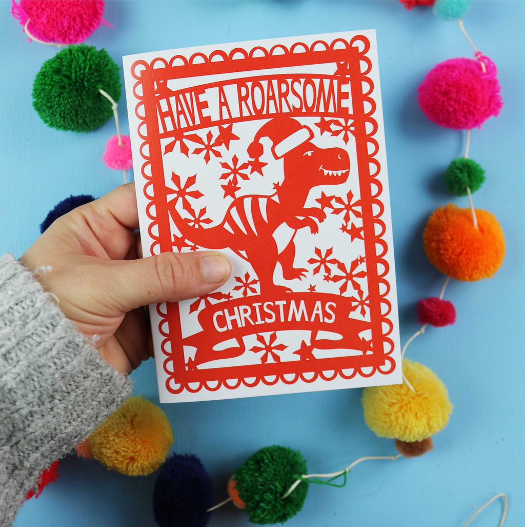 Hand holding a Christmas card with a dinosaur design against a colourful pom-pom background