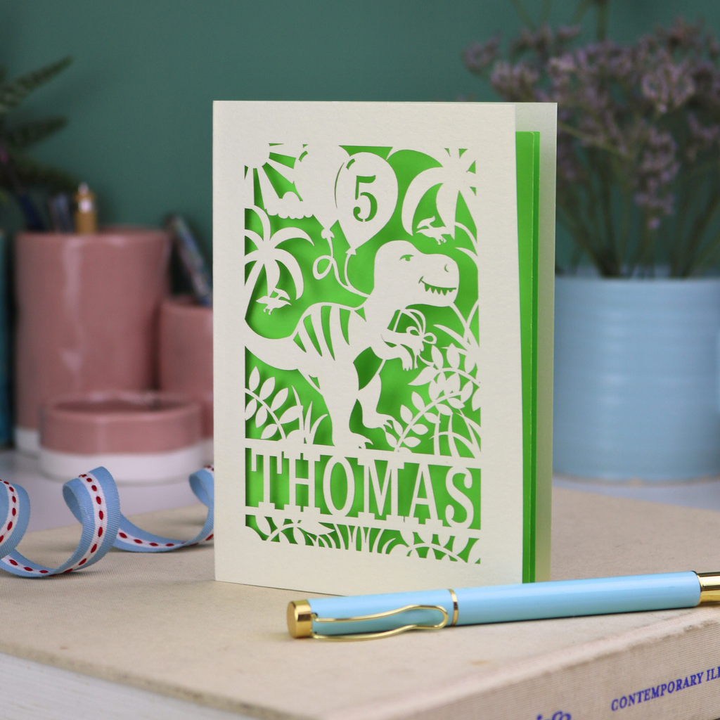 A laser cut dinosaur birthday card, personalised with a cut out name across the bottom. The birthday card features a papercut illustration of a dinosaur holding 2 balloons, one of them has a cut out number to show the age of the recipient.  