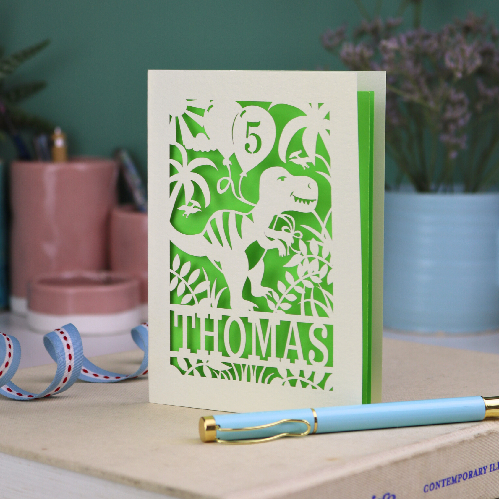 A laser cut dinosaur birthday card, personalised with a cut out name across the bottom. The birthday card features a papercut illustration of a dinosaur holding 2 balloons, one of them has a cut out number to show the age of the recipient.  