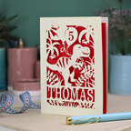 A laser cut dinosaur birthday card, personalised with a cut out name across the bottom. The birthday card features a papercut illustration of a dinosaur holding 2 balloons, one of them has a cut out number to show the age of the recipient.  