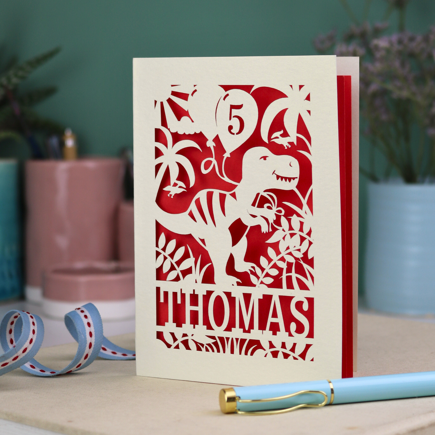 A laser cut dinosaur birthday card, personalised with a cut out name across the bottom. The birthday card features a papercut illustration of a dinosaur holding 2 balloons, one of them has a cut out number to show the age of the recipient.  