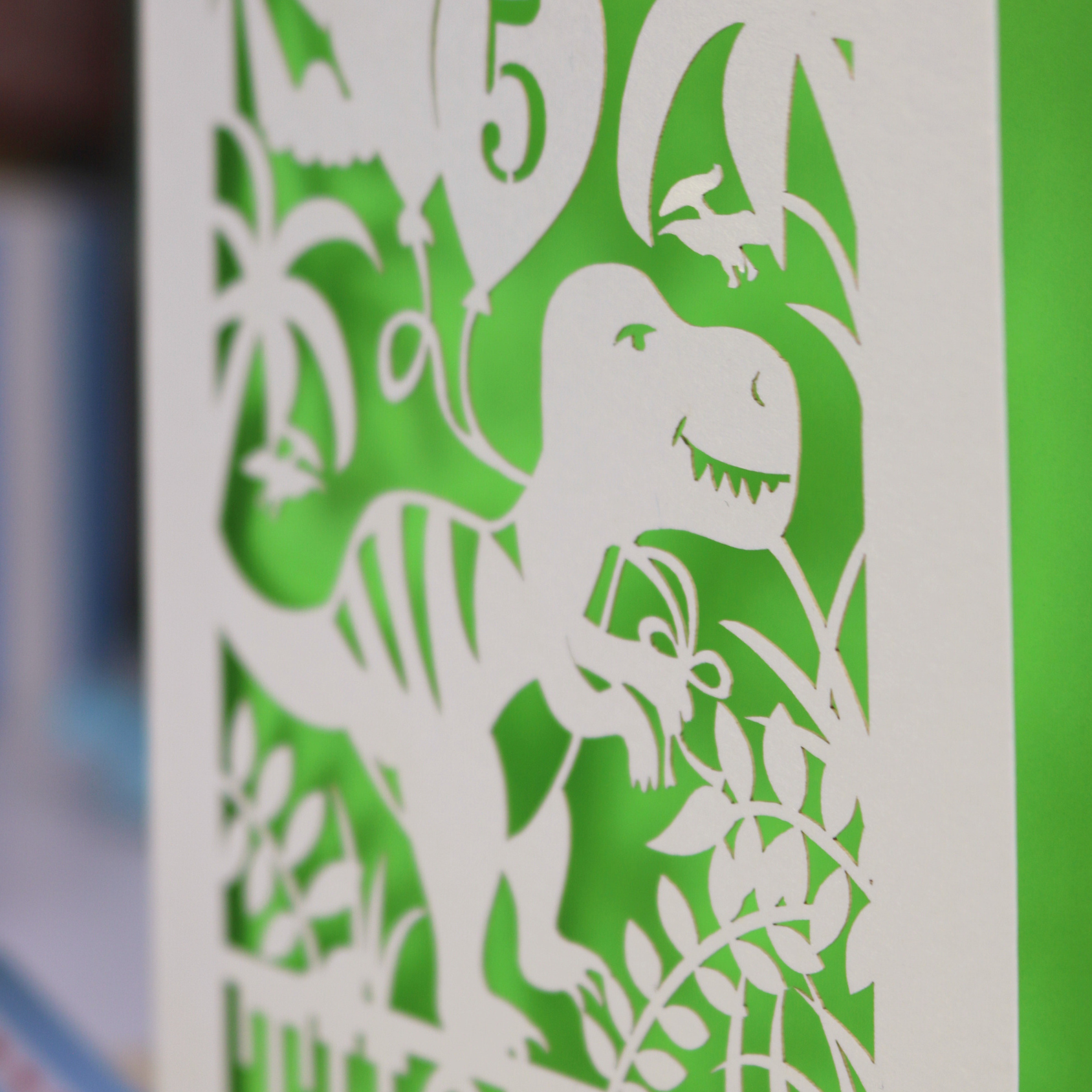 A laser cut dinosaur birthday card, personalised with a cut out name across the bottom. The birthday card features a papercut illustration of a dinosaur holding 2 balloons, one of them has a cut out number to show the age of the recipient.  