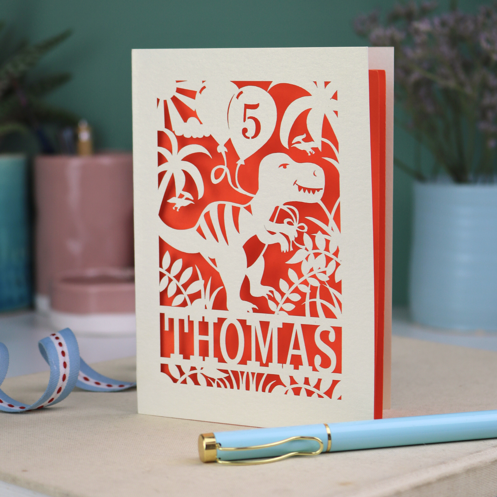 A laser cut dinosaur birthday card, personalised with a cut out name across the bottom. The birthday card features a papercut illustration of a dinosaur holding 2 balloons, one of them has a cut out number to show the age of the recipient.  