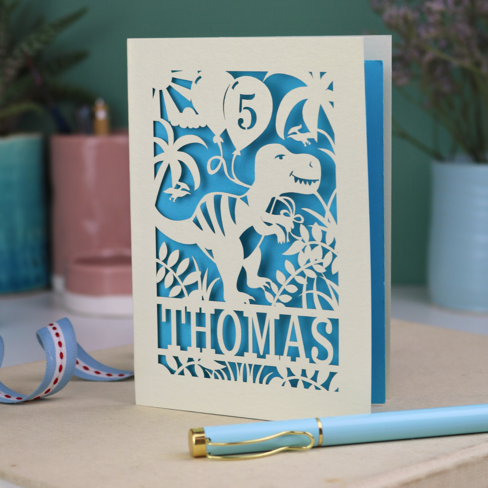A laser cut dinosaur birthday card, personalised with a cut out name across the bottom. The birthday card features a papercut illustration of a dinosaur holding 2 balloons, one of them has a cut out number to show the age of the recipient.  
