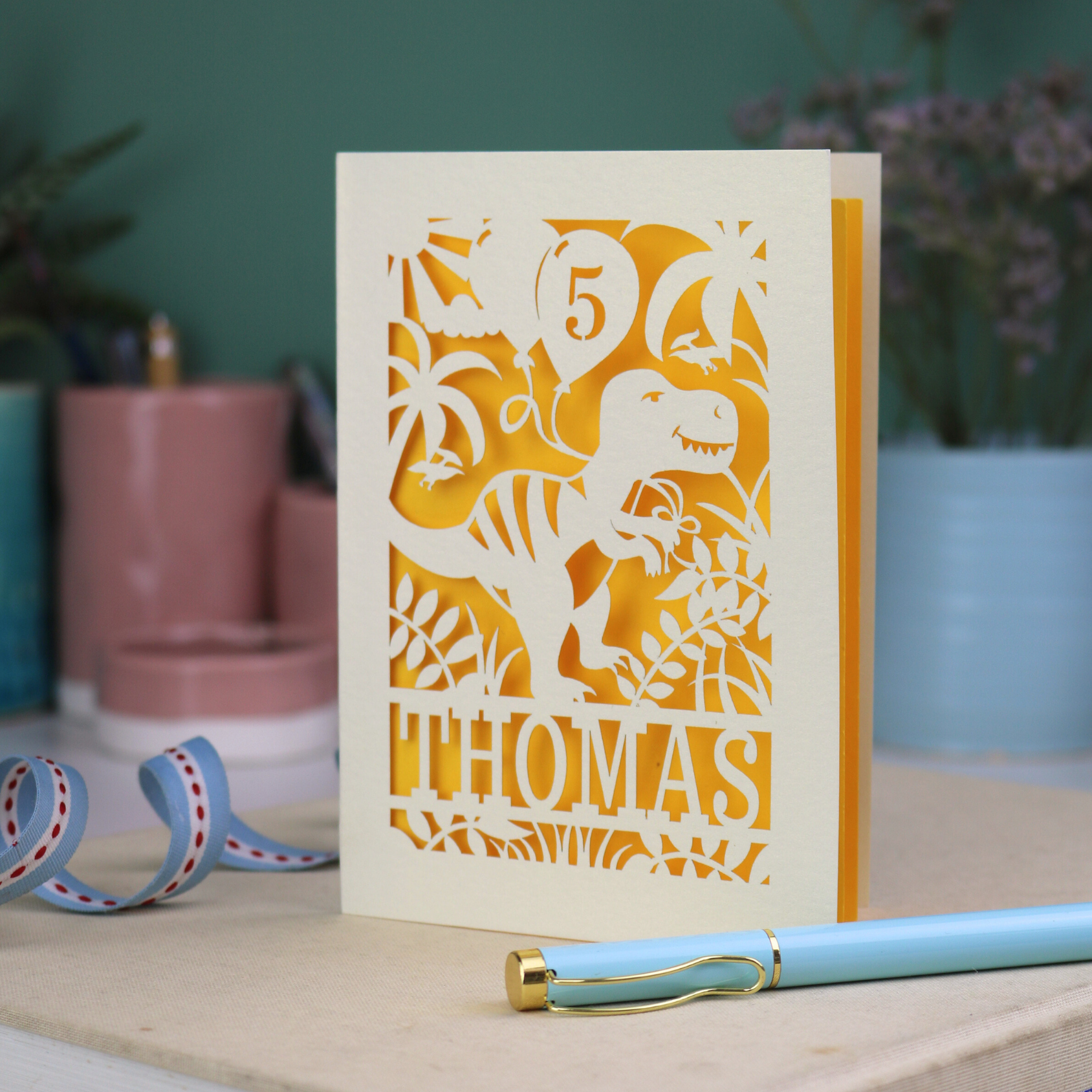 A laser cut dinosaur birthday card, personalised with a cut out name across the bottom. The birthday card features a papercut illustration of a dinosaur holding 2 balloons, one of them has a cut out number to show the age of the recipient.  