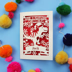 Laser cut Christmas card with dinosaur design and colourful pom-poms on a blue background