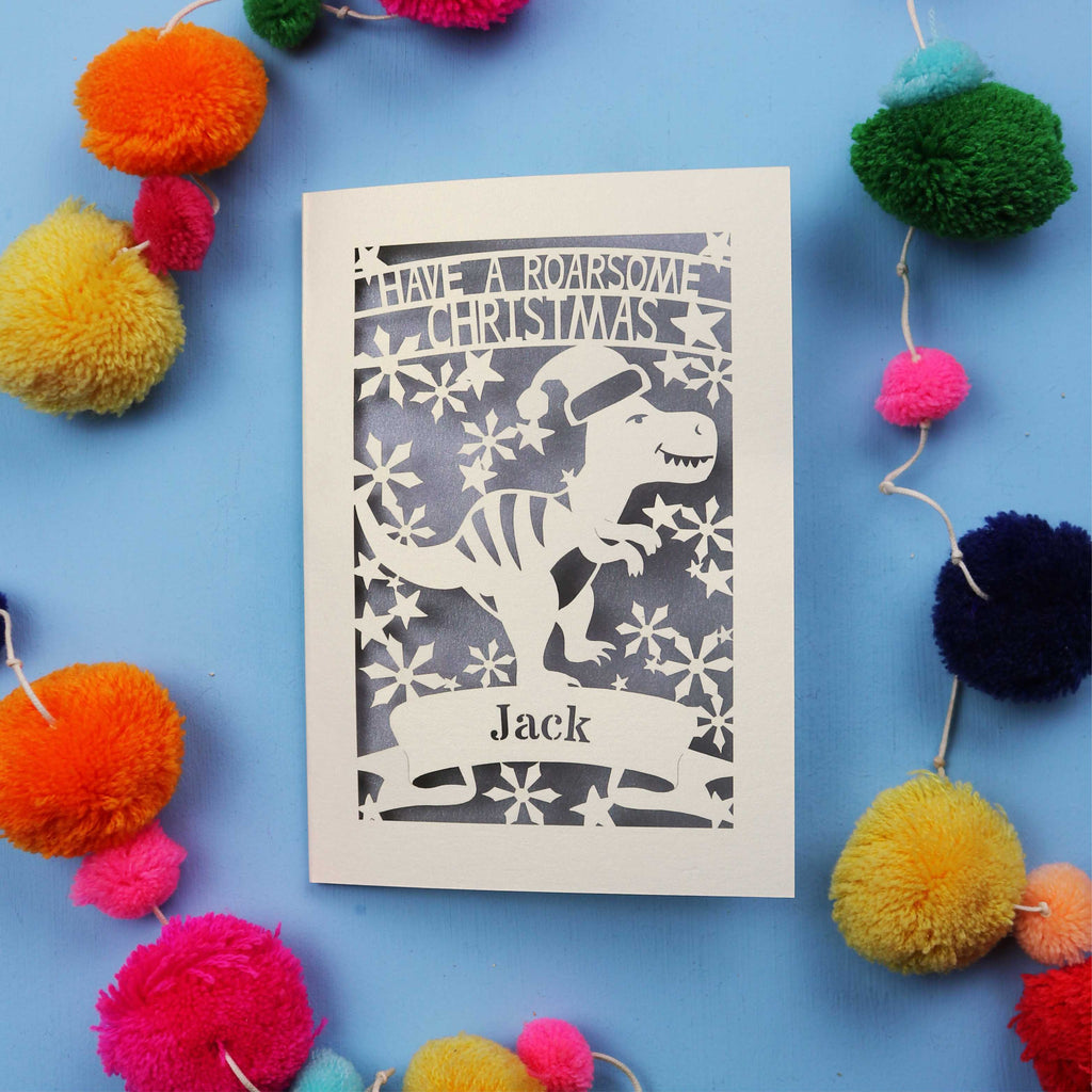 Laser cut Christmas card with dinosaur design and colourful pom-poms on a blue background