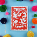 Red Christmas card with dinosaur design on a blue background with colourful pom-poms.
