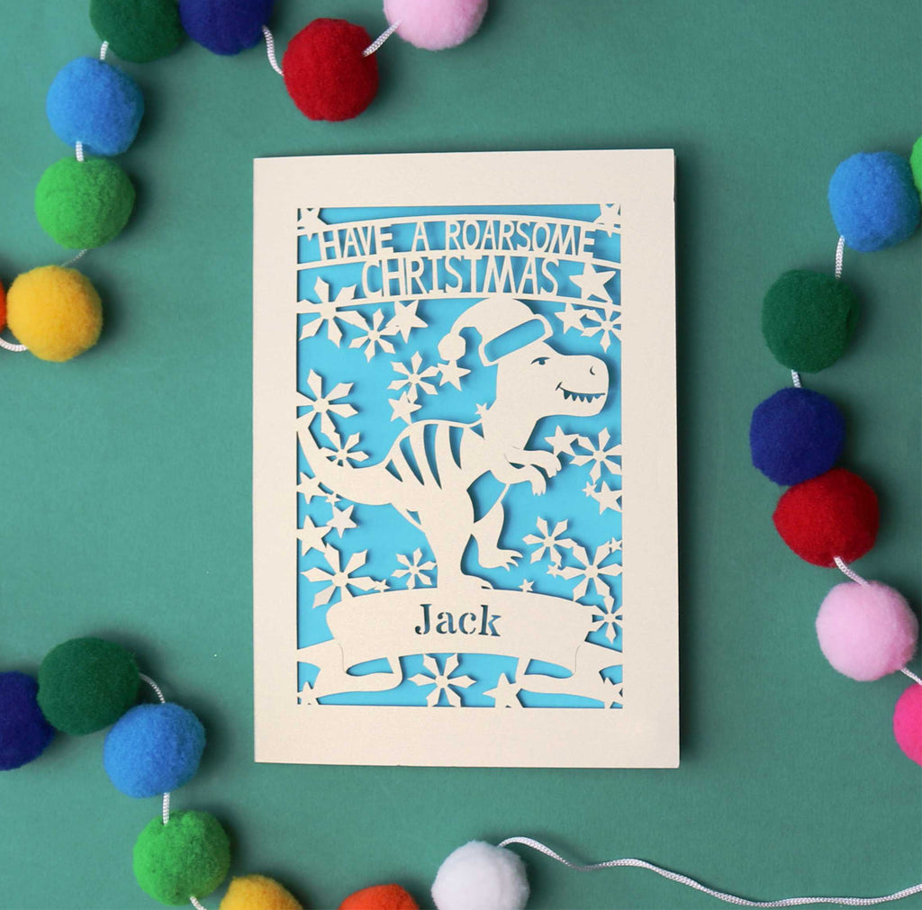 Laser cut Christmas card with dinosaur design and colourful pom-poms on a green background