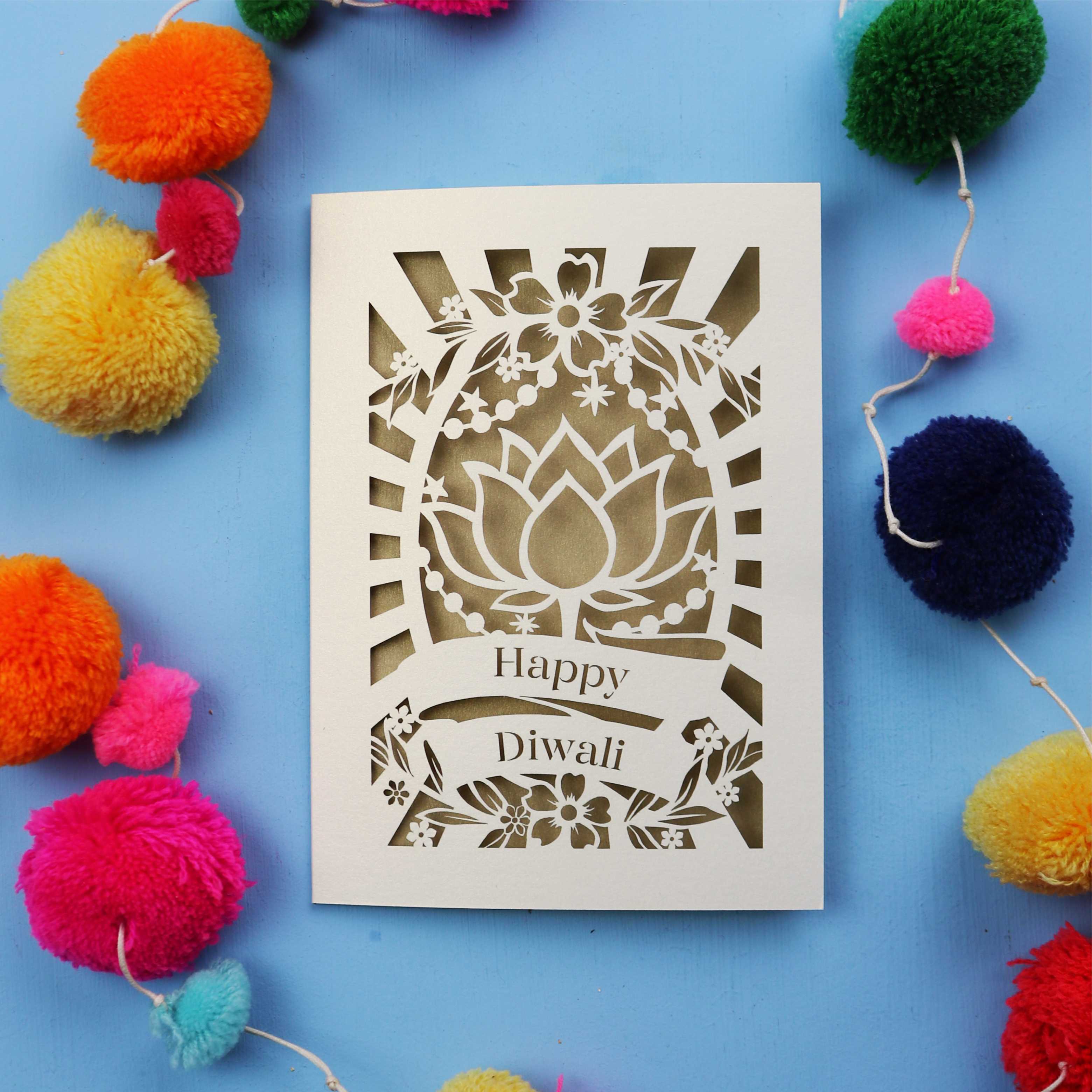 Decorative laser cut Diwali card with 'Happy Diwali' text on a blue background with colourful pom-poms.