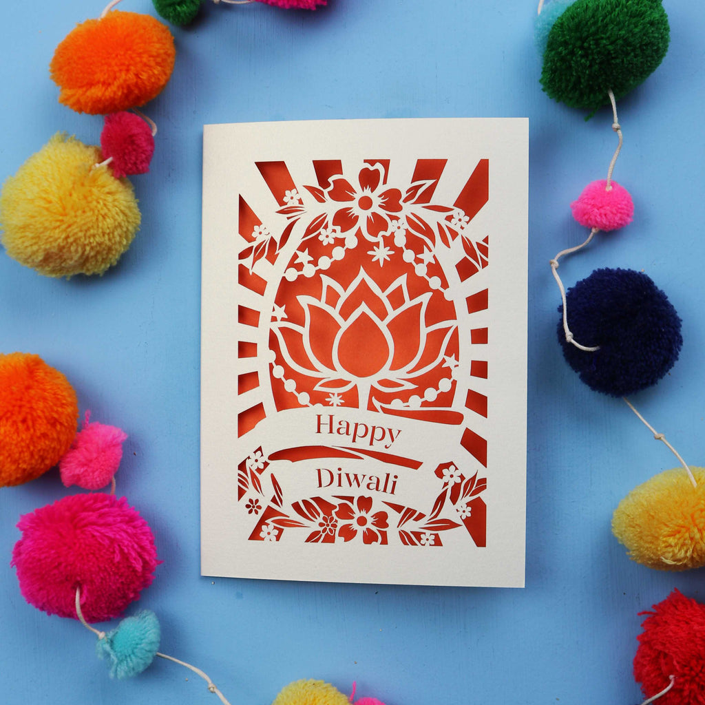 Decorative laser cut Diwali card with 'Happy Diwali' text on a blue background with colourful pom-poms.