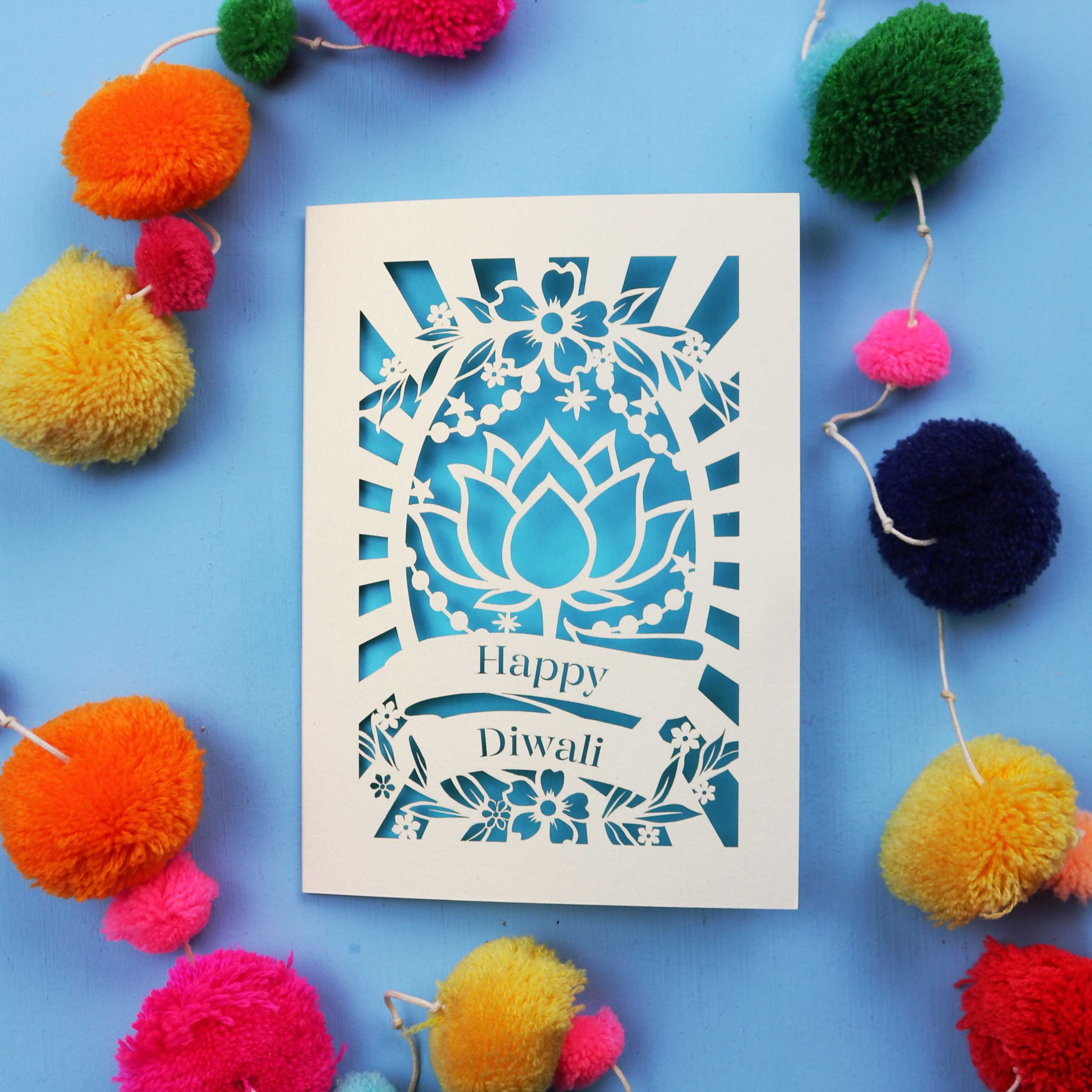 Decorative laser cut Diwali card with 'Happy Diwali' text on a blue background with colourful pom-poms.