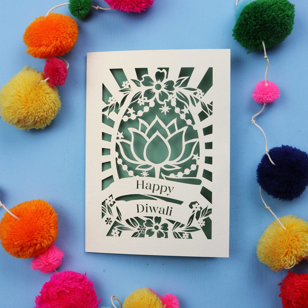Decorative laser cut Diwali card with 'Happy Diwali' text on a blue background with colourful pom-poms.