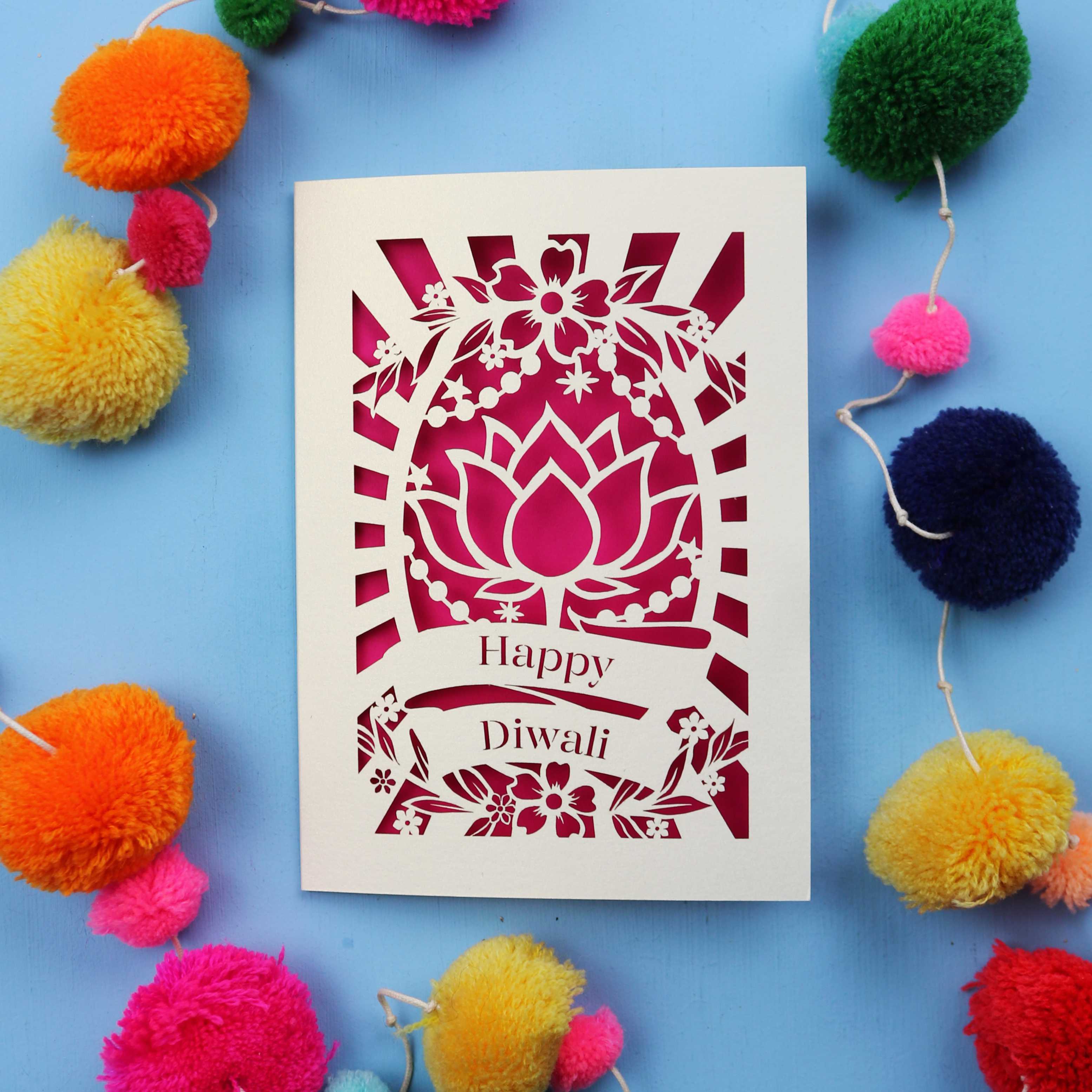 Decorative laser cut Diwali card with 'Happy Diwali' text on a blue background with colourful pom-poms.