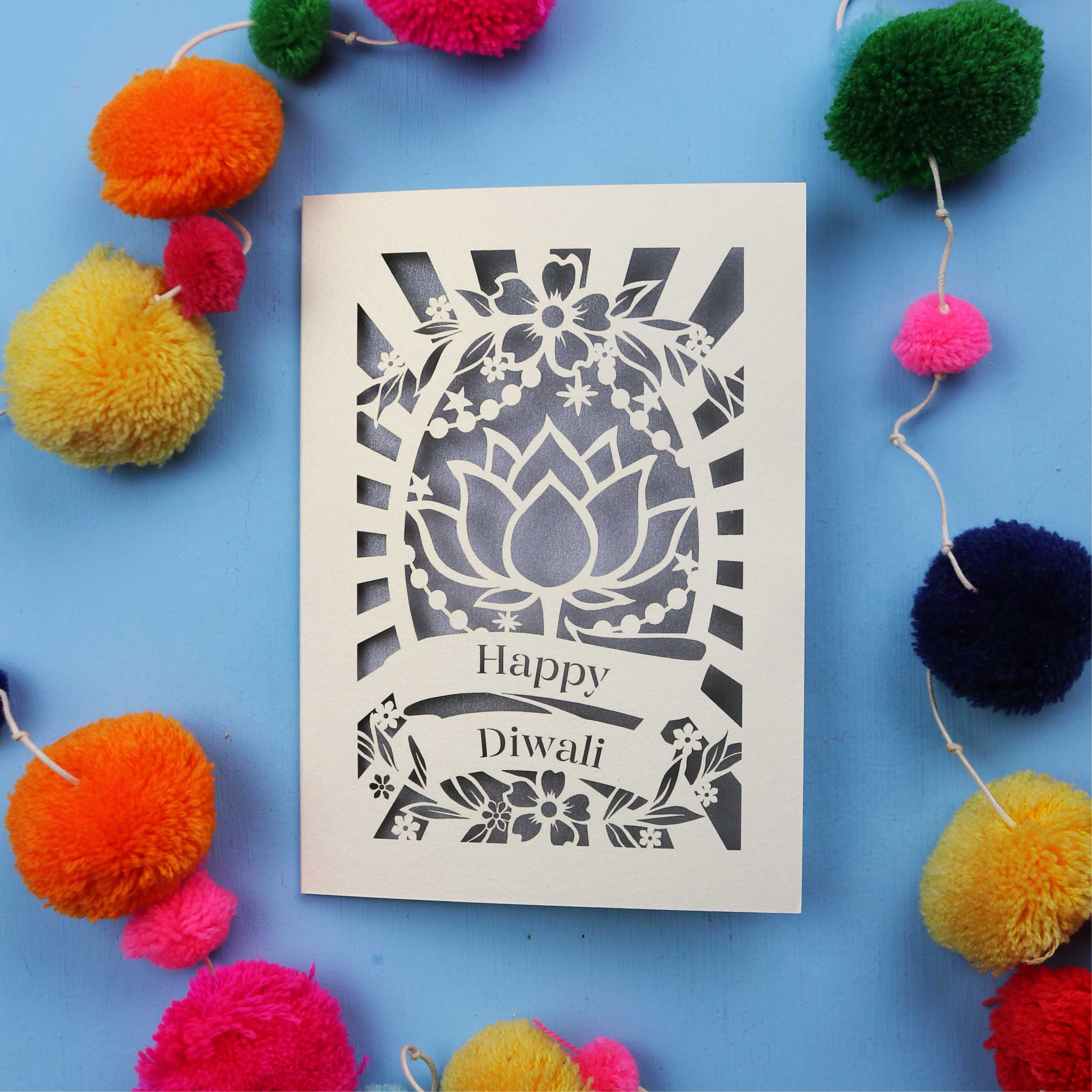 Decorative laser cut Diwali card with 'Happy Diwali' text on a blue background with colourful pom-poms.