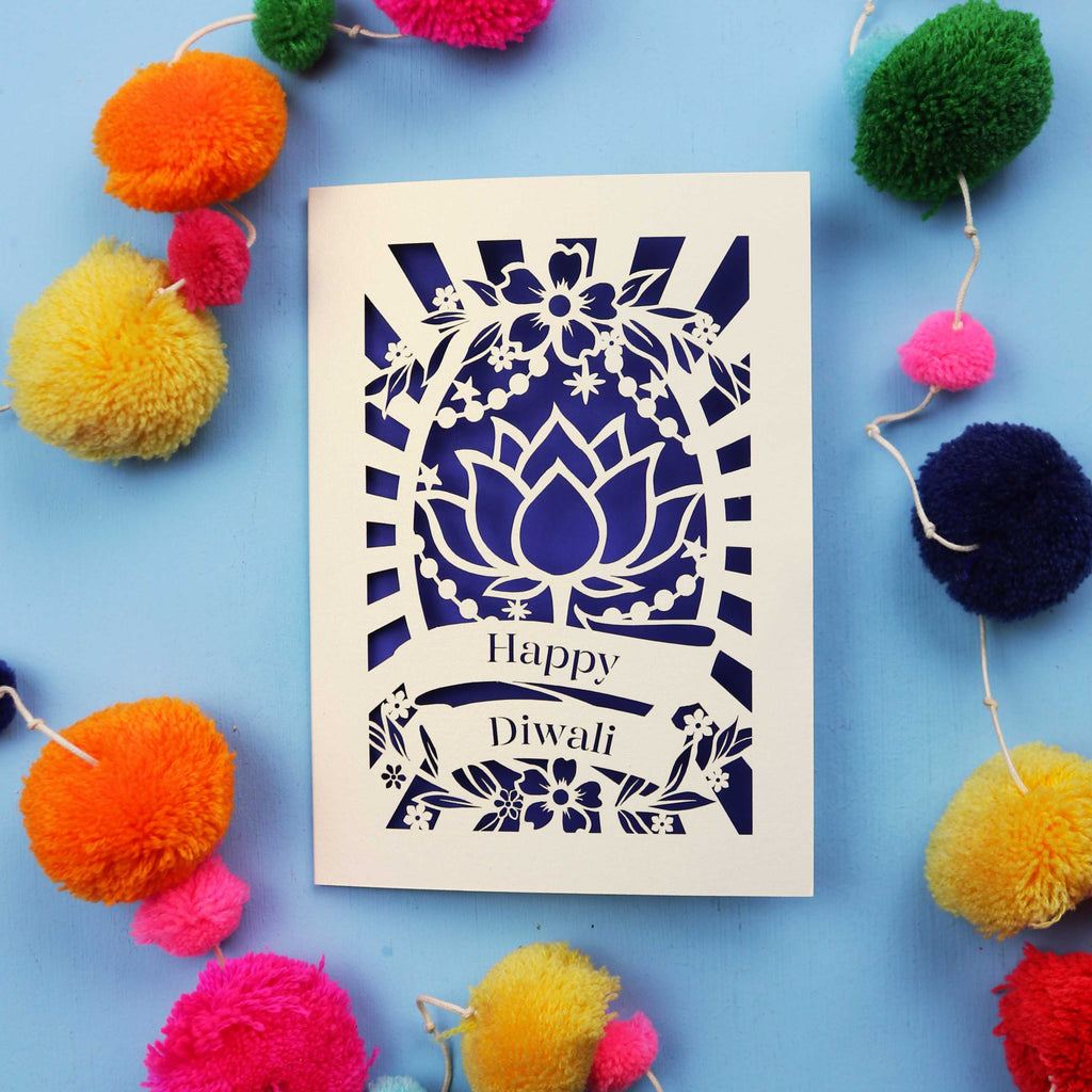 Decorative laser cut Diwali card with 'Happy Diwali' text on a blue background with colourful pom-poms.