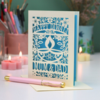 A decorative greeting card with intricate laser-cut floral and diya designs, reading 'HAPPY DIWALI 2025 MUM & DAD,' displayed on a table with candles, a pink pen, and stationery in the background.