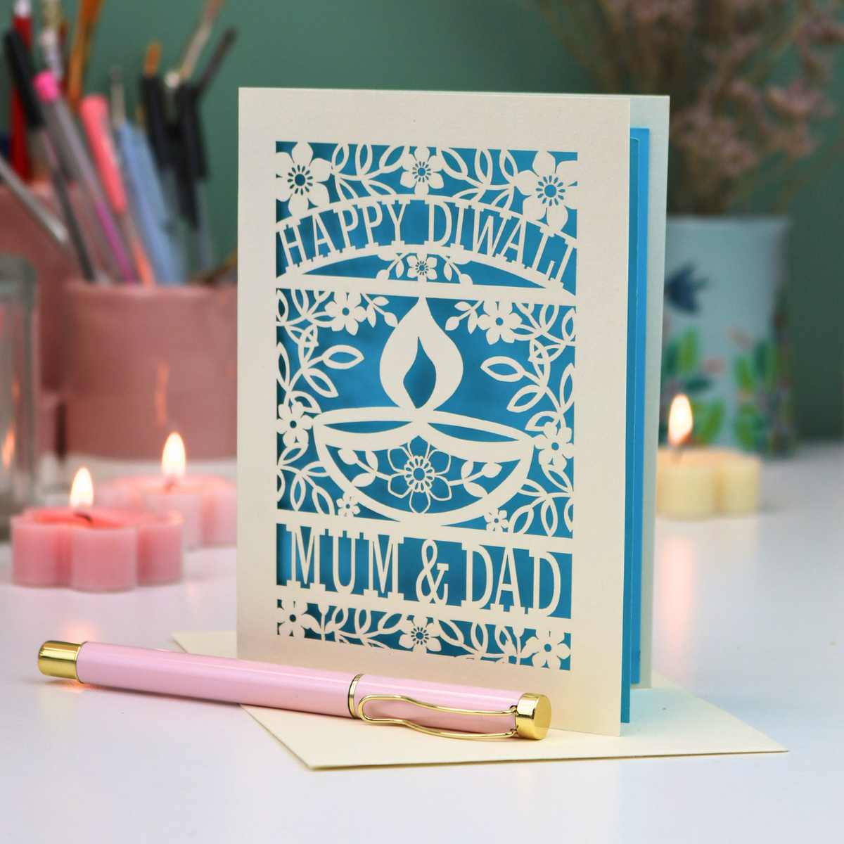 A decorative greeting card with intricate laser-cut floral and diya designs, reading 'HAPPY DIWALI 2025 MUM & DAD,' displayed on a table with candles, a pink pen, and stationery in the background.
