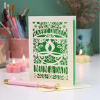 A cream-colored laser-cut Diwali greeting card with floral and diya designs, reading 'HAPPY DIWALI' and 'MUM & DAD', displayed on a table with candles and a pink pen in a softly lit setting.