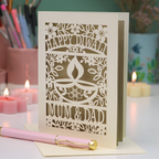 A cream-colored laser-cut Diwali greeting card with floral and diya designs, reading 'HAPPY DIWALI' and 'MUM & DAD', displayed on a table with candles and a pink pen in a softly lit setting.