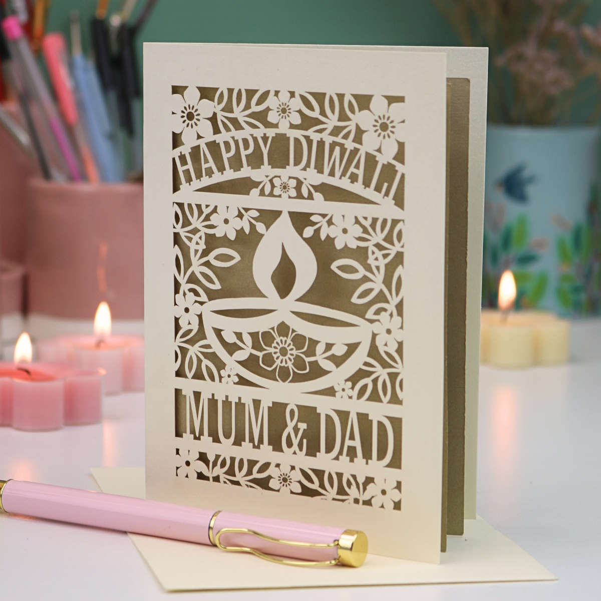 A cream-colored laser-cut Diwali greeting card with floral and diya designs, reading 'HAPPY DIWALI' and 'MUM & DAD', displayed on a table with candles and a pink pen in a softly lit setting.