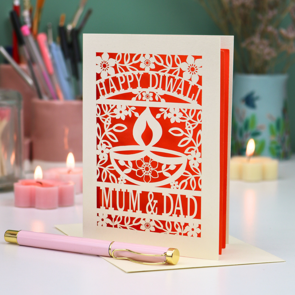 A cream-colored laser-cut Diwali greeting card with floral and diya designs, reading 'HAPPY DIWALI' and 'MUM & DAD', displayed on a table with candles and a pink pen in a softly lit setting.