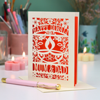 A cream-colored laser-cut Diwali greeting card with floral and diya designs, reading 'HAPPY DIWALI' and 'MUM & DAD', displayed on a table with candles and a pink pen in a softly lit setting.