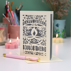 A cream-colored laser-cut Diwali greeting card with floral and diya design, reading 'HAPPY DIWALI' and 'MUM & DAD', displayed on a table with candles and a pink pen in a softly lit setting.