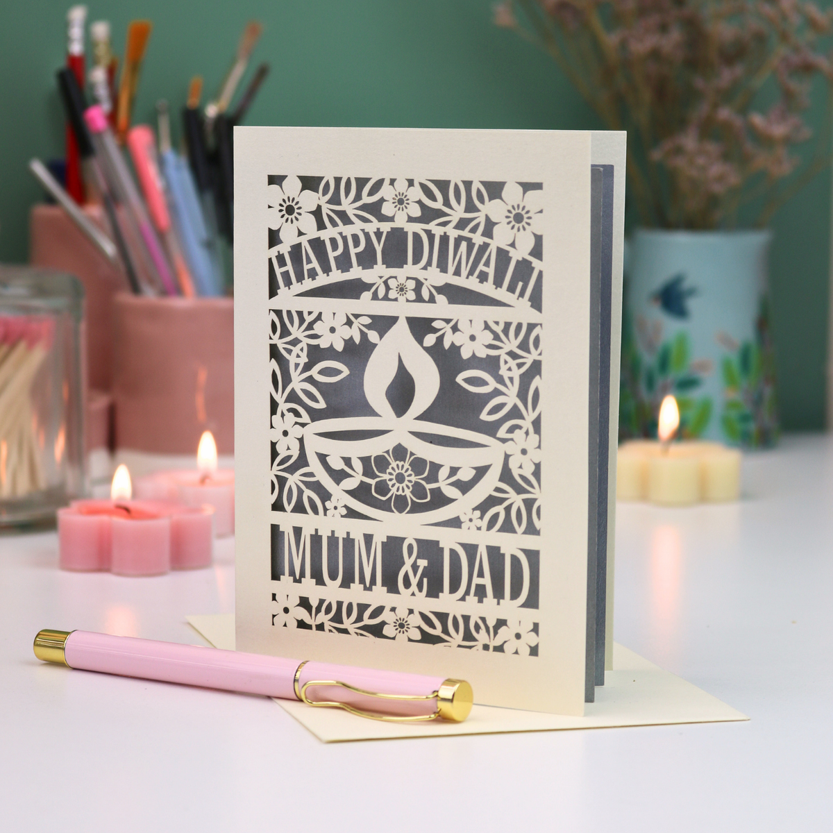 A cream-colored laser-cut Diwali greeting card with floral and diya design, reading 'HAPPY DIWALI' and 'MUM & DAD', displayed on a table with candles and a pink pen in a softly lit setting.