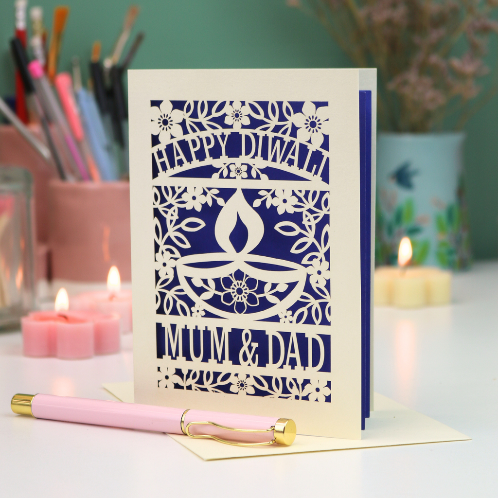 A cream-colored laser-cut Diwali greeting card with floral and diya designs, reading 'HAPPY DIWALI' and 'MUM & DAD', displayed on a table with candles and a pink pen in a softly lit setting.