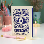 A cream-colored laser-cut Diwali greeting card with floral and diya designs, reading 'HAPPY DIWALI' and 'MUM & DAD', displayed on a table with candles and a pink pen in a softly lit setting.
