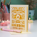 A handmade laser-cut Diwali greeting card for 'Mum & Dad' standing on a white surface with a pink pen resting in front. The card is cream-colored with intricate cutouts showing a yellow interior, featuring a diya and floral patterns.