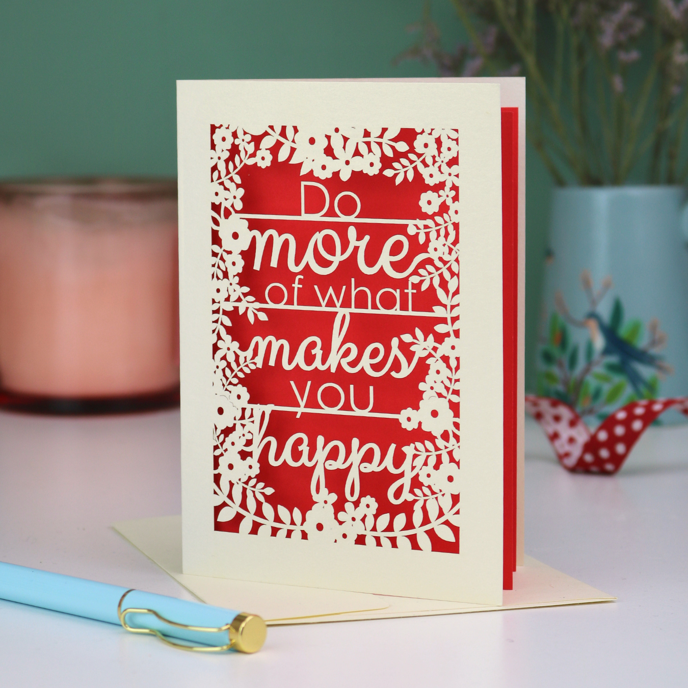 Motivational card with text 'Do more of what makes you happy' on a red background, placed on a surface with a pen and decorative items.