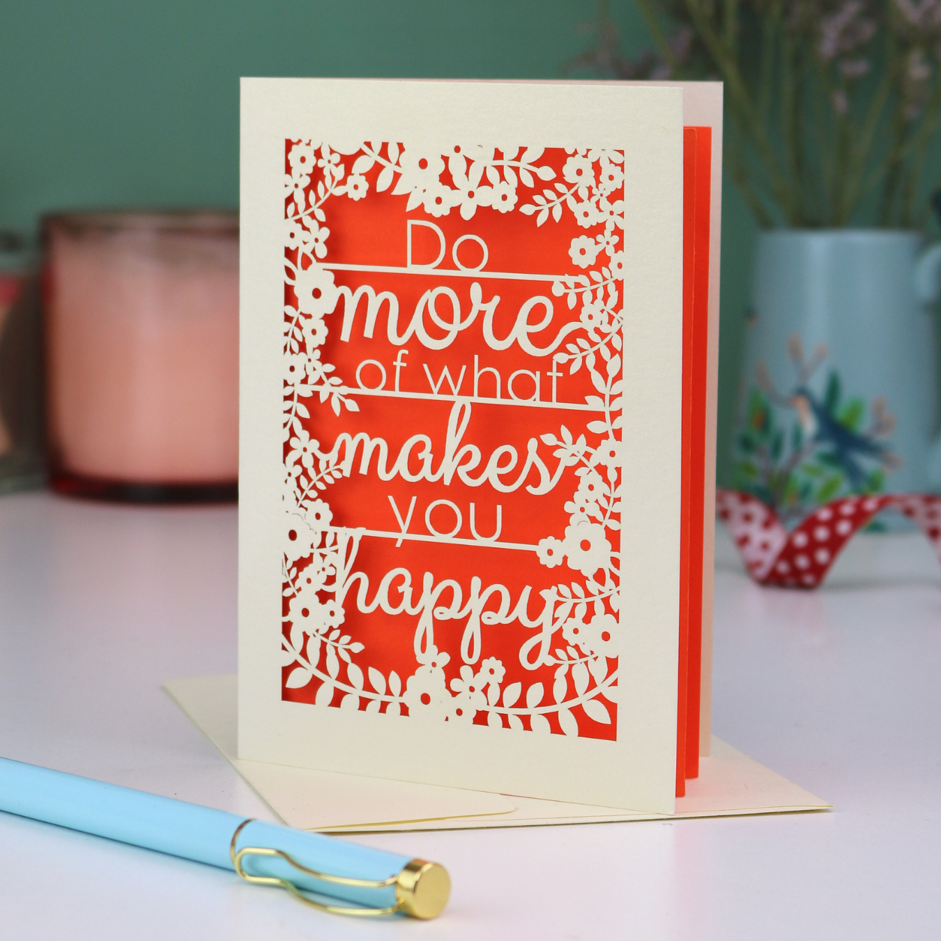 Greeting card with motivational quote, "Do more of what makes you happy" on a table with a pen and decorative items.