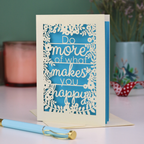 Laser cut inspirational quote card with text 'Do more of what makes you happy' on a blue background, placed on a surface with a pen and decorative items.