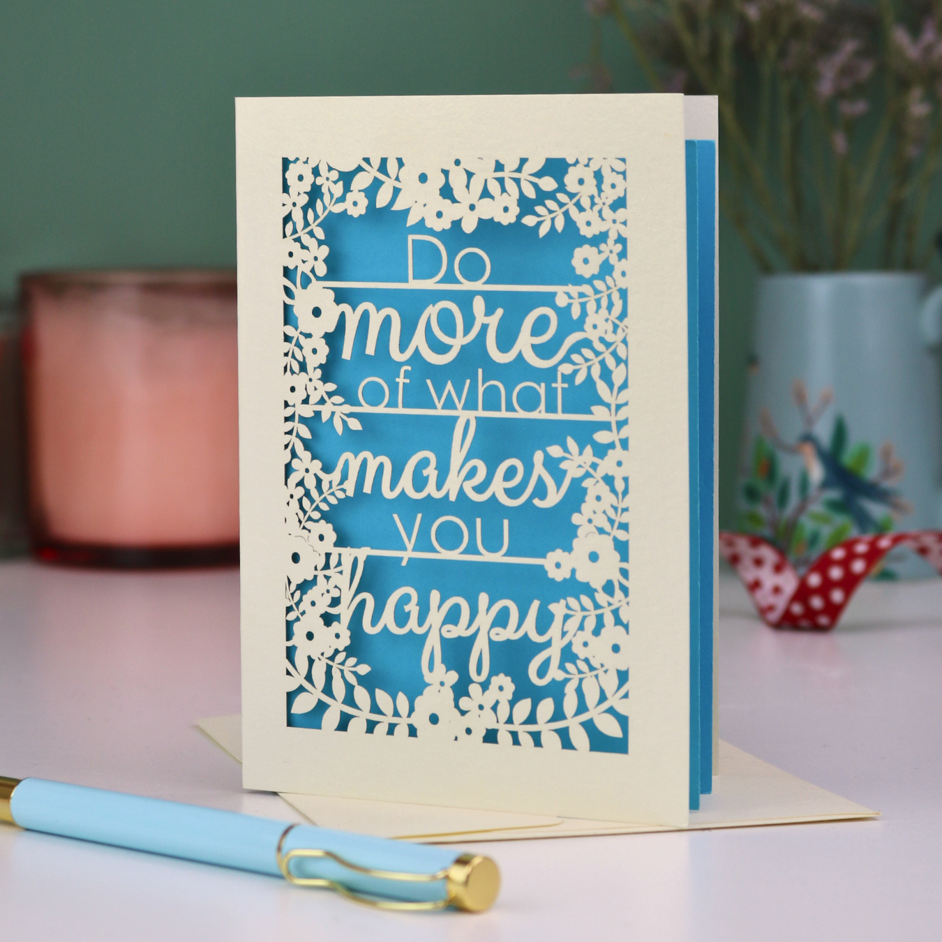 Laser cut inspirational quote card with text 'Do more of what makes you happy' on a blue background, placed on a surface with a pen and decorative items.