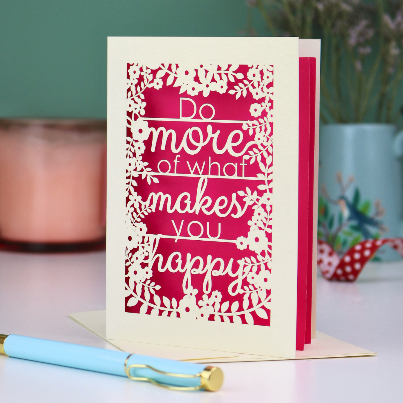 Laser cut inspirational card with text 'Do more of what makes you happy' on a pink background, placed on a surface with a pen and decorative items.