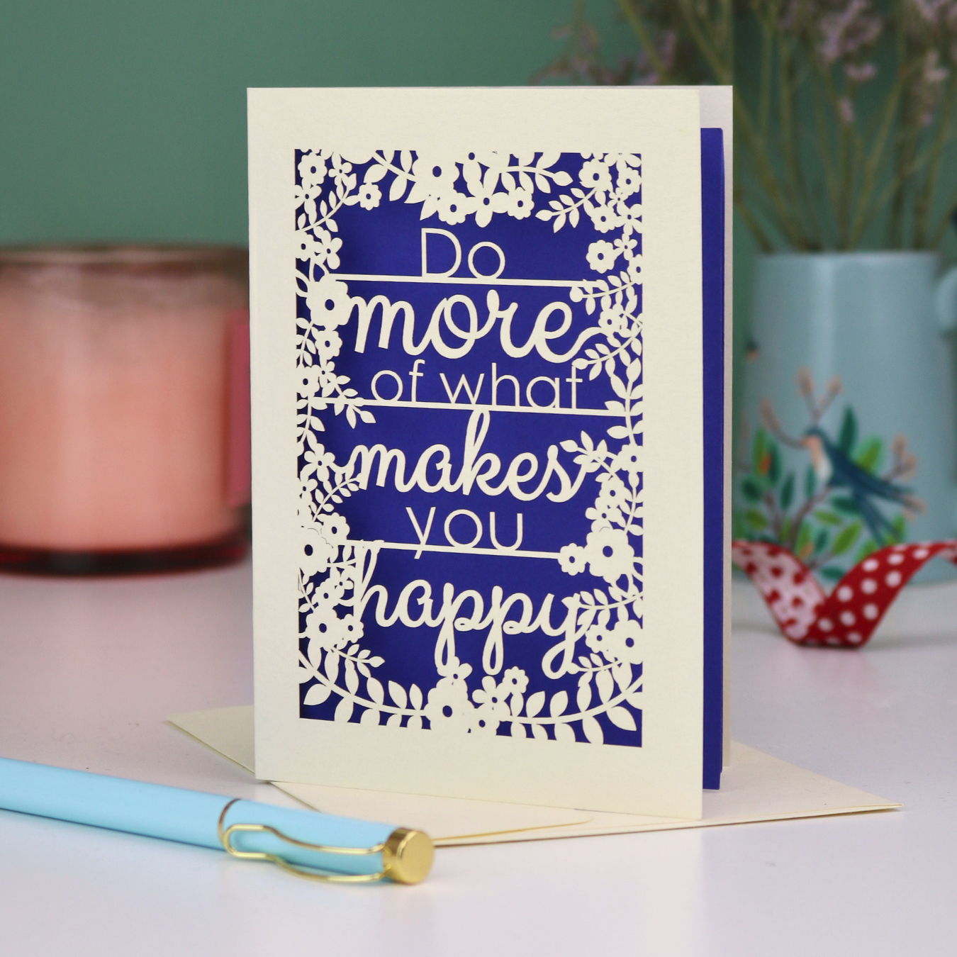 Laser cut encouragement card with text 'Do more of what makes you happy' on a violet background, placed on a surface with a pen and decorative items.