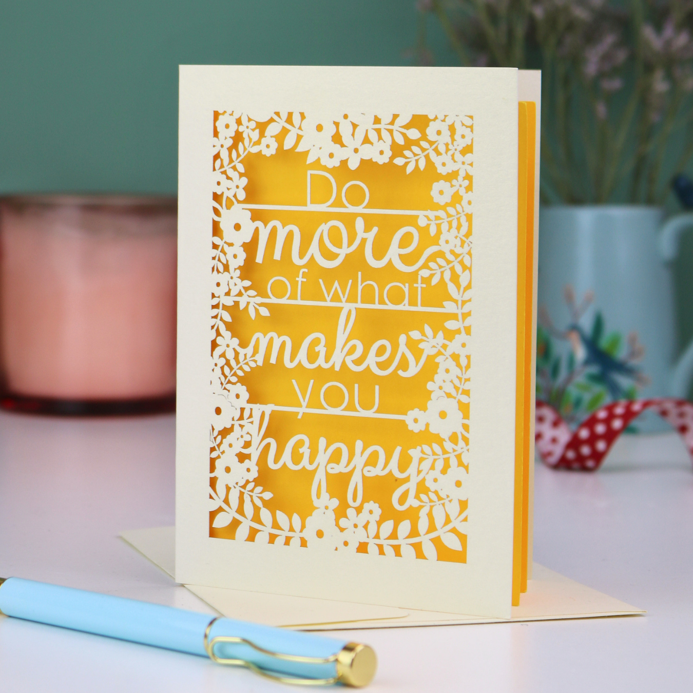 Laser cut Motivational card with text 'Do more of what makes you happy' on a yellow background, placed on a surface with a pen and decorative items.