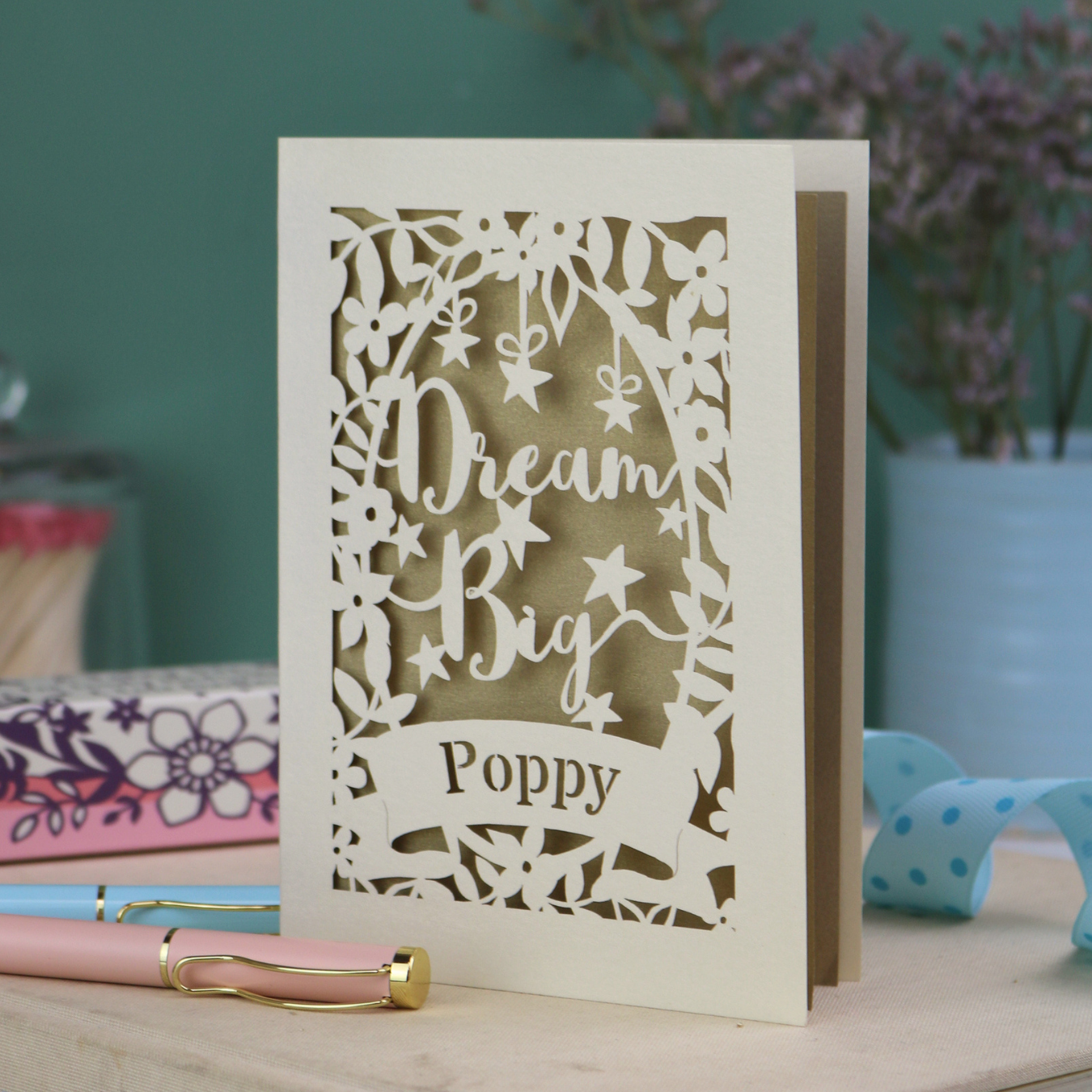 Laser-cut good luck card with 'Dream Big Poppy' text on a wooden surface with stationery items.