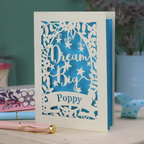 Personalised good luck Card with 'Dream Big Poppy' text on a blue and cream design, placed on a surface with stationery items.