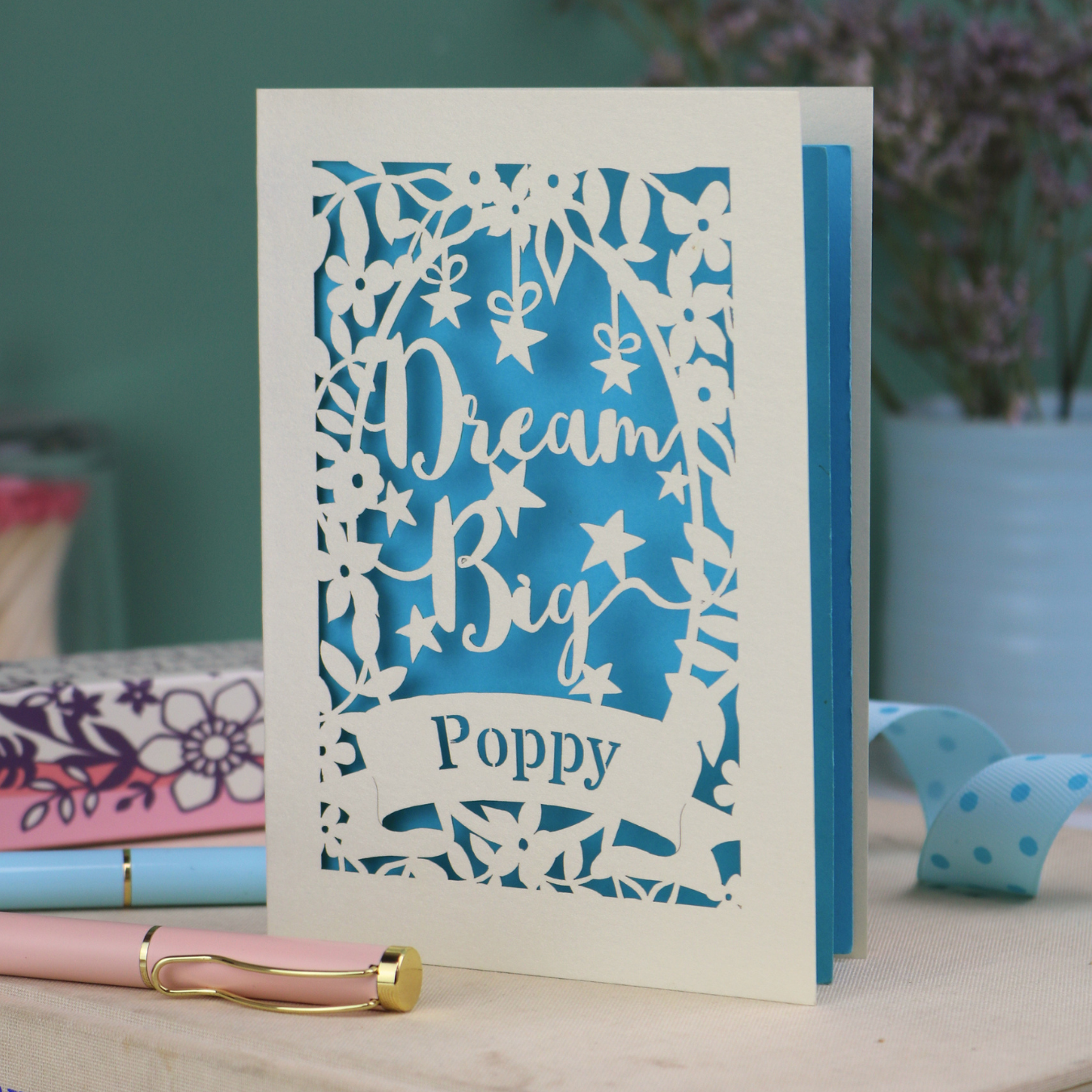 Personalised good luck Card with 'Dream Big Poppy' text on a blue and cream design, placed on a surface with stationery items.