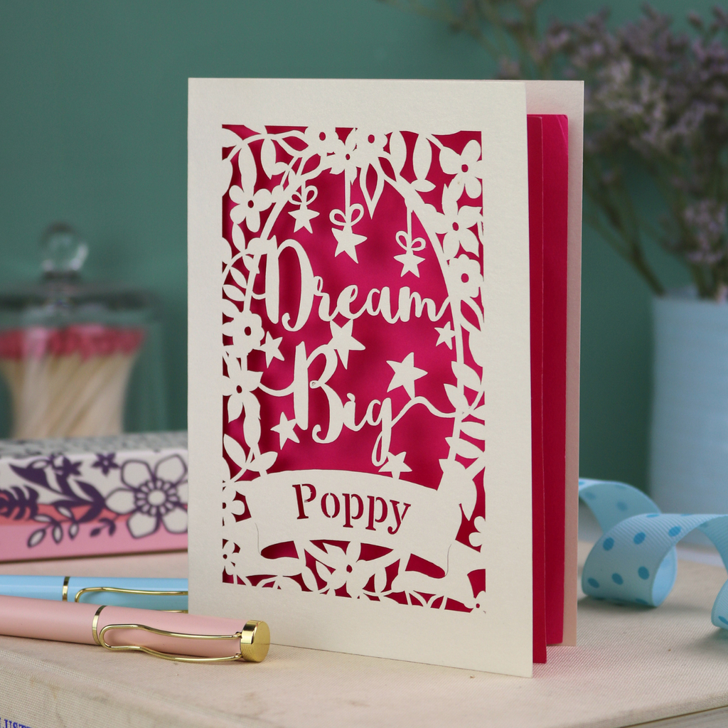 Greeting card for good luck with 'Dream Big Poppy' text on a desk with decorative items.