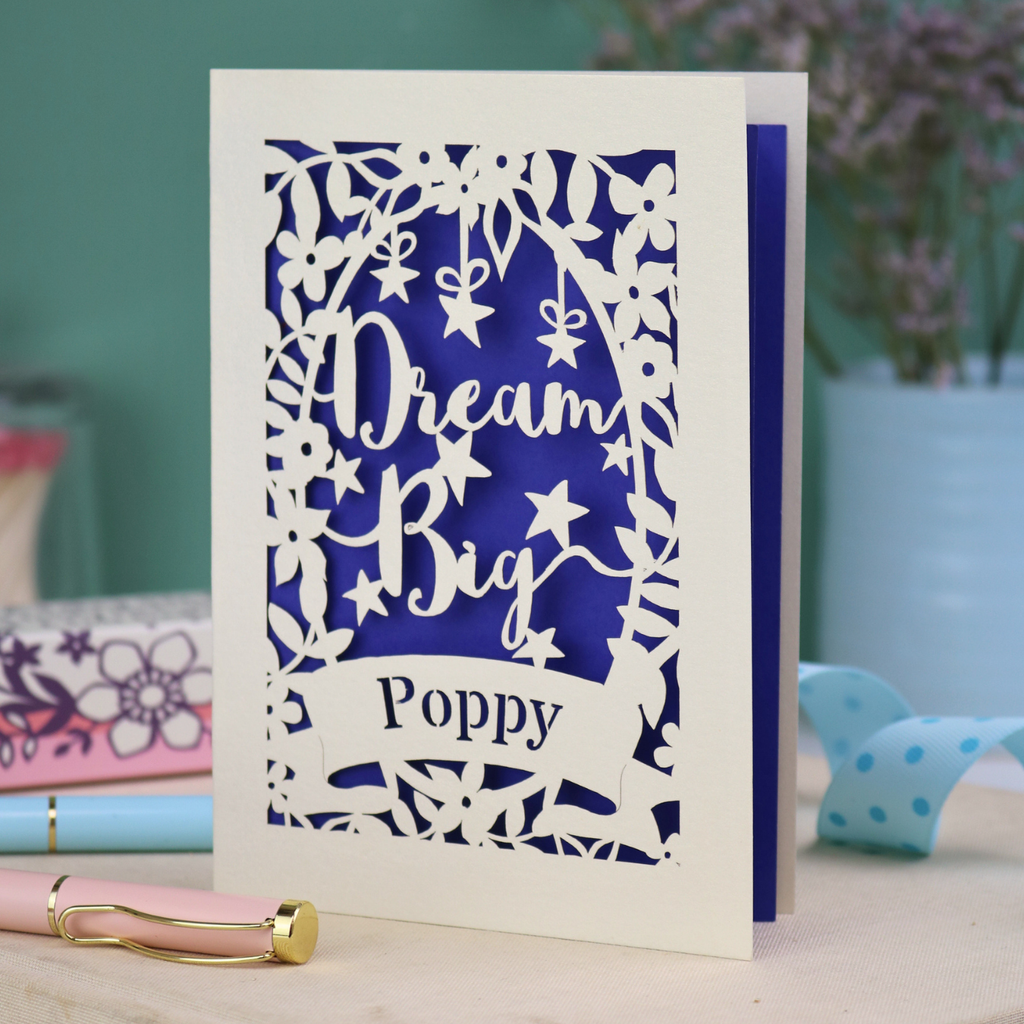 Laser cut good luck Card with 'Dream Big Poppy' text on a blue and white design, placed on a surface with a blurred background.