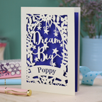 Laser cut good luck Card with 'Dream Big Poppy' text on a blue and white design, placed on a surface with a blurred background.