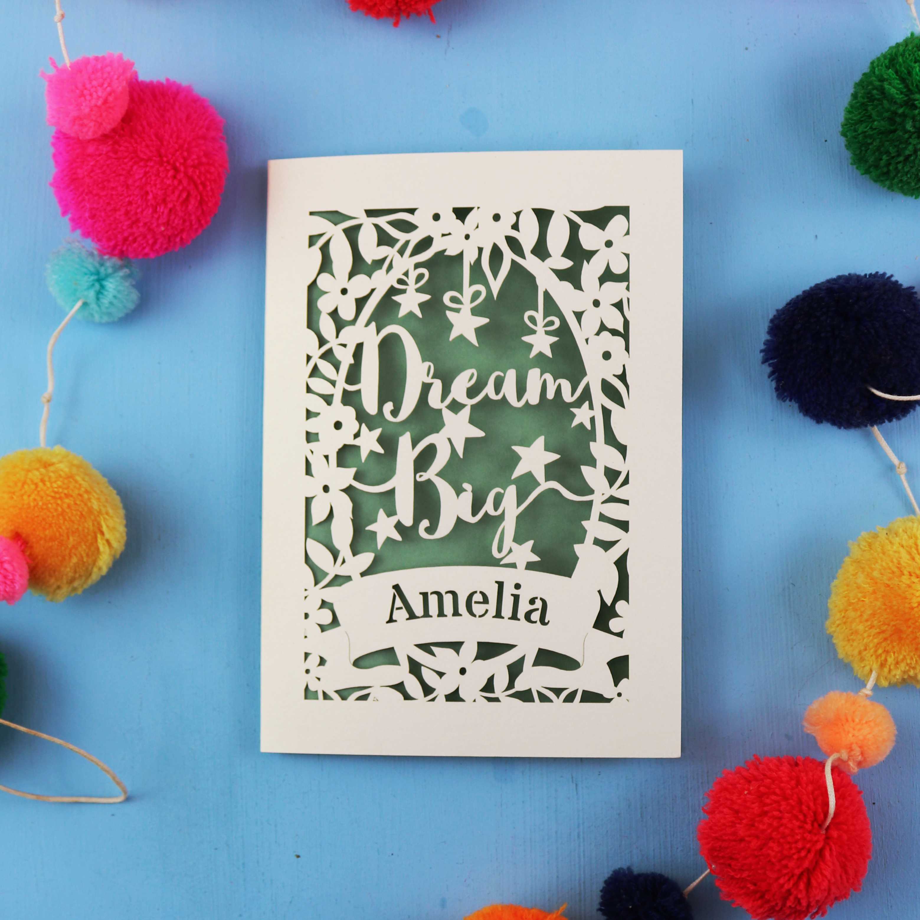 Decorative card with 'Dream Big Amelia' text on a blue background with colourful pom-poms.
