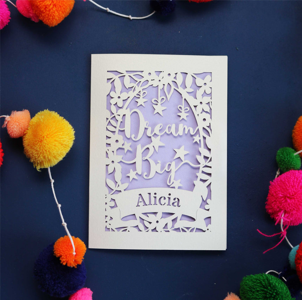 A personalised laser cut good luck card with the words 'Dream Big' and a name 'Alicia' written on it, displayed on a dark background surrounded by colourful pompons.