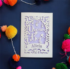 A personalised laser cut good luck card with the words 'Dream Big' and a name 'Alicia' written on it, displayed on a dark background surrounded by colourful pompons.