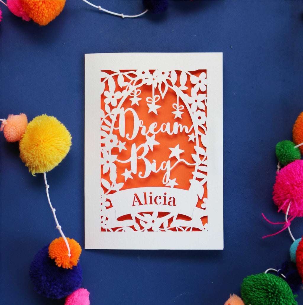 A personalised laser cut card with the words 'Dream Big' and a name 'Alicia' written on it, displayed on a dark background surrounded by colourful pompons.