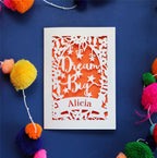 A personalised laser cut card with the words 'Dream Big' and a name 'Alicia' written on it, displayed on a dark background surrounded by colourful pompons.